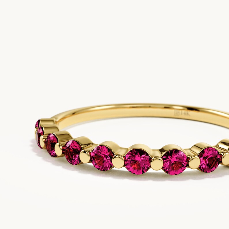 May include: A yellow gold band ring featuring a series of round, vibrant pink gemstones set in a row. The metal band is polished and smooth, with the engraving 14K visible on the inner surface of the ring, showcasing a classic design with stones.