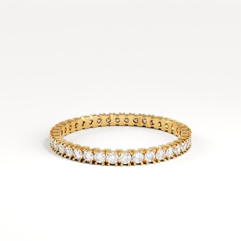 May include: A gold eternity ring with a continuous row of round, clear gemstones. The ring is set against a white background, highlighting the sparkle of the gemstones and the warm tone of the gold.