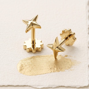 May include: A pair of gold-toned stud earrings, each featuring a four-pointed star design. The earrings have a flat, flower-shaped backing and a smooth, polished metallic finish, presented with a gold paint stroke on a textured surface.