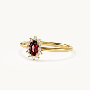 May include: A gold ring featuring a central oval-cut deep red garnet gemstone surrounded by a halo of small, round, clear white diamonds. The band is a polished yellow gold, creating a classic look with a vibrant contrast between the red stone.