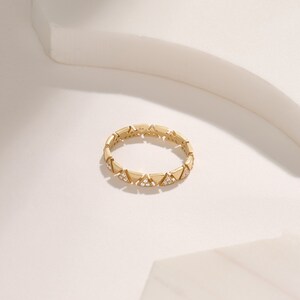 May include: A gold ring featuring a repeating triangular pattern. Each triangle is embellished with small, clear gemstones. The ring is displayed on a white surface, accentuating its intricate design and elegant style.