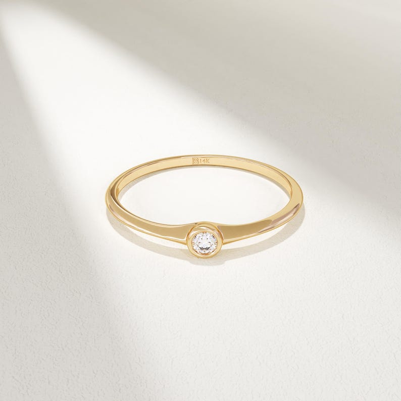 May include: A delicate gold ring with a single, round, clear gemstone set in the center. The ring band is thin and smooth, with a slight widening at the gemstone setting. The ring is displayed on a white surface.