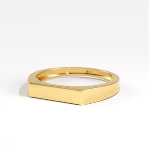 Op de afbeelding: Een gouden ring met een rechthoekige voorkant en afgeronde randen. De ring is gemaakt van gepolijst goud en heeft een glad, reflecterend oppervlak. De ring staat tegen een witte achtergrond.