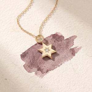 Könnte beinhalten: Eine goldene Halskette mit einem sechsstrahligen Sternanhänger, der in der Mitte mit einem kleinen, klaren, rund geschliffenen Edelstein besetzt ist. Der Anhänger hängt an einer zarten Goldkette und hat eine polierte Metalloberfläche.