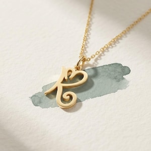 May include: A gold-toned necklace featuring a delicate chain and a pendant shaped like the letter K, designed with a stylized, cursive-inspired font that incorporates a heart-like loop at the top and a decorative swirl at the base.