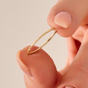 May include: A delicate gold ring with a twisted rope design. The ring is held between fingers, showcasing its slender profile and intricate texture. The ring is marked with "14K".