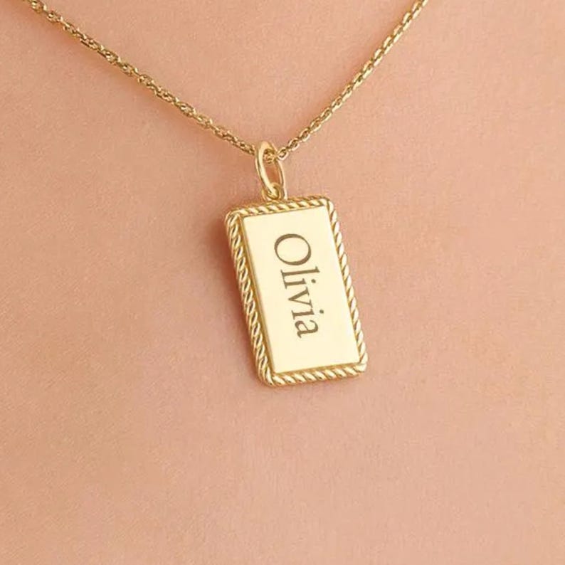 May include: A gold-toned necklace featuring a rectangular pendant with a rope-style border. The centre of the pendant is smooth and engraved with the name Olivia in a classic serif font. The pendant hangs from a delicate gold-toned chain.