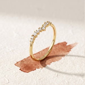 May include: A gold ring featuring a delicate, V-shaped chevron band set with seven small, round, clear stones along the top curve. The metal has a polished finish, and the interior of the band is clearly stamped with the text 14K.