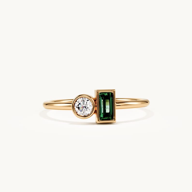 May include: A gold ring featuring a round, bezel-set clear gemstone next to a rectangular, emerald-green baguette-cut gemstone. The band is smooth and polished, showcasing a minimalist design that highlights the contrast between the two stones.