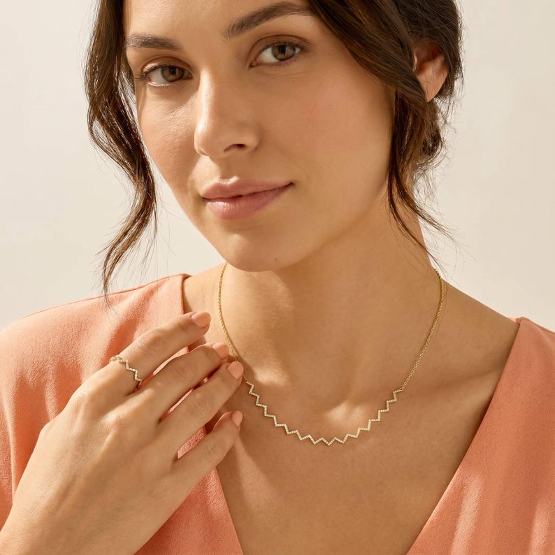 May include: A gold necklace and ring set. The necklace features a zigzag design, and the ring has a matching pattern. The jewelry is worn on a person with a peach-colored top.