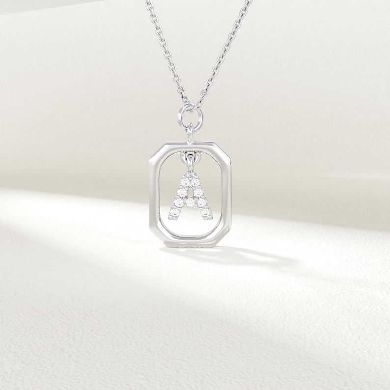 May include: Silver pendant necklace with the letter "A" formed from clear gemstones. The letter is set within a rectangular frame, suspended from a fine chain. The jewellery is presented against a soft, neutral background.