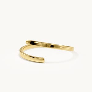Könnte beinhalten: Ein polierter goldfarbener Bypass-Ring mit einem glatten, durchgehenden Band, das sich in einem offenen, überlappenden Design um sich selbst wickelt. Die Metalloberfläche ist stark reflektierend und zeigt ein helles, metallisch gelbes Finish.