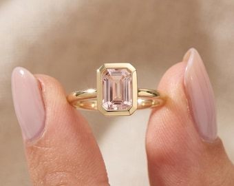 Könnte beinhalten: Ein Goldring mit einem rechteckigen, im Smaragdschliff geschliffenen, hellrosa Edelstein in einer glatten Zargenfassung. Die Ringschiene ist aus poliertem Gold gefertigt und der Stein ist mittig platziert, mit einer klaren, facettierten Oberfläche.