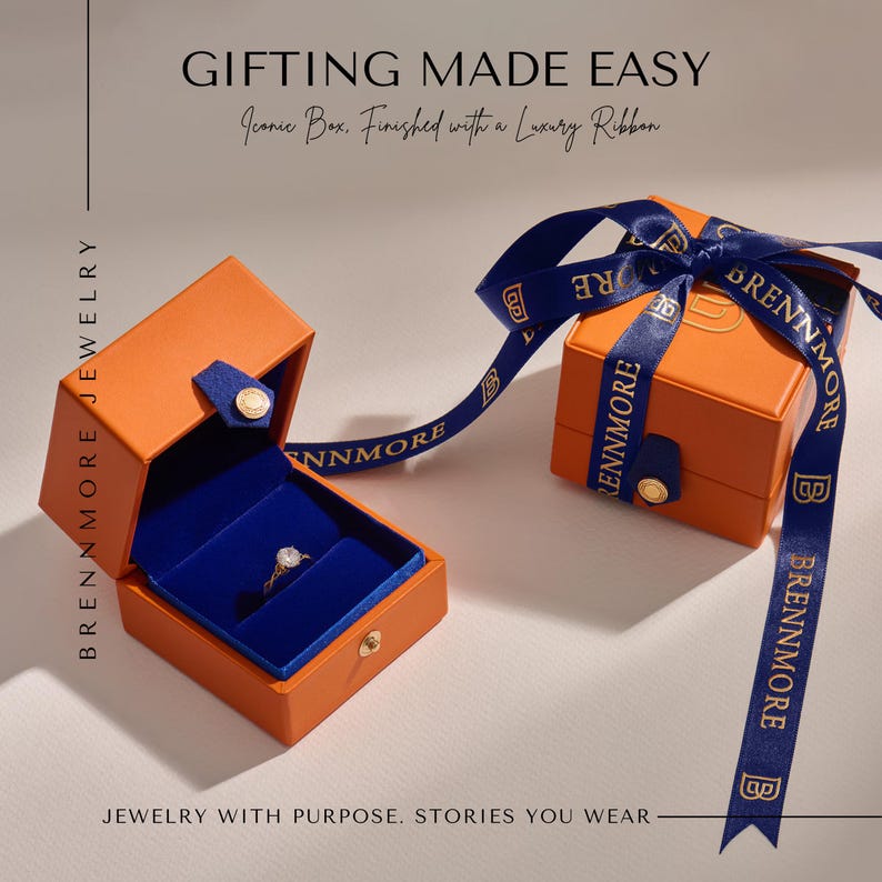 May include: Two orange jewelry boxes with blue velvet interiors and gold-tone snap closures. One box is open, displaying a gold ring with a clear stone. The closed box is tied with a blue ribbon featuring the text BRENNMORE. Text reads GIFTING MADE EASY.
