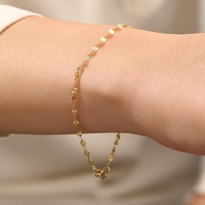 May include: A delicate gold chain bracelet featuring a series of small, circular, textured metal discs linked together. The bracelet has a polished finish and includes a small metal clasp at the end, creating a refined and simple jewelry piece.