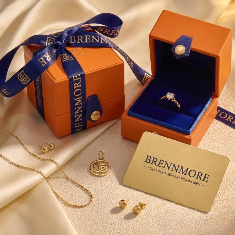 Op de afbeelding: Een oranje geschenkdoos met een blauw lint en een bijpassende open doos met een diamanten ring. Gouden sieraden, waaronder een ketting, oorbellen en een hanger, zijn ook aanwezig. Een gouden kaart leest "BRENNMORE - SOLID GOLD JEWELRY FOR WOMEN."