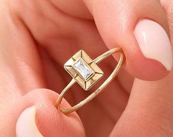 May include: A gold ring featuring a rectangular baguette-cut clear gemstone set in a geometric gold frame. The thin gold band is stamped with the text 14K on the inner surface, indicating the metal purity of the jewelry piece.