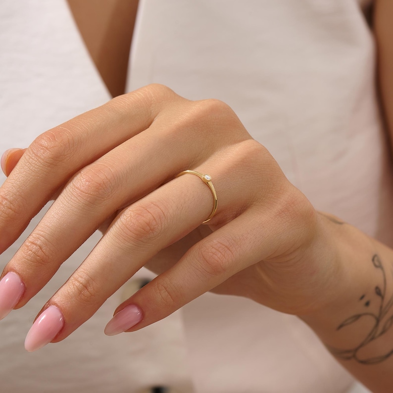 May include: A delicate gold ring with a small, clear gemstone. The ring is thin and minimalist, featuring a single, round stone set in a simple band. The ring is worn on a finger, showcasing its elegant design and subtle sparkle.
