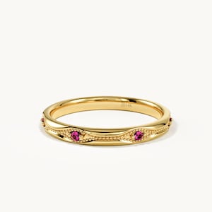 May include: A gold band ring featuring a delicate beaded texture along the outer edge and two small, round, vibrant pink gemstones set into the metal. The interior of the band is clearly stamped with the text 14K to indicate the metal purity.