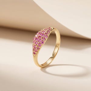 Op de afbeelding: Een gouden ring met een gebogen band, bezet met talrijke kleine roze edelstenen. De ring heeft een gepolijste afwerking en wordt gepresenteerd tegen een neutrale achtergrond. De edelstenen zijn dicht op elkaar geplaatst, waardoor een effen roze oppervlak ontstaat.