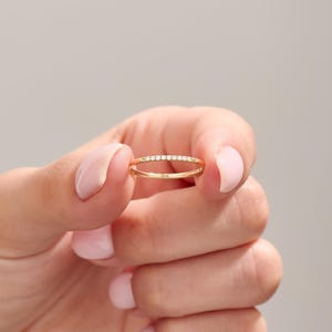 May include: A delicate gold ring with a row of small, clear gemstones set across the top. The ring is held between fingers against a soft gray background. The ring is a simple band design.