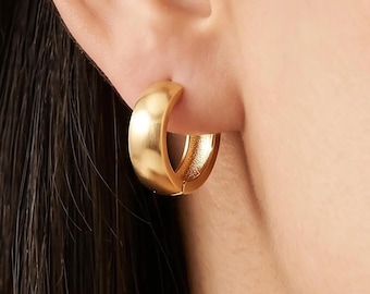 May include: A thick, polished gold hoop earring with a smooth, rounded surface and a hinged closure mechanism. The earring features a high-shine metallic finish that reflects light, creating a bright, warm glow against the earlobe.