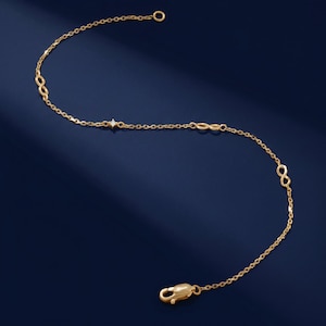 Könnte beinhalten: Ein zartes goldfarbenes Armband mit Unendlichkeitszeichen und einem kleinen Diamanten. Das Armband hat einen Karabinerverschluss und einen kleinen Ring. Der Schmuck wird auf einem dunkelblauen Hintergrund präsentiert.