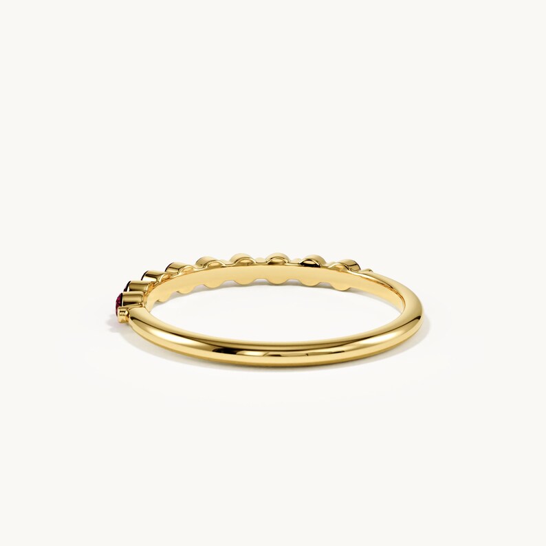 May include: A polished yellow gold ring featuring a series of small, round, deep red gemstones set along the top half of the band. The band has a smooth, rounded profile and a high-shine metallic finish, showcasing a minimalist and elegant design.
