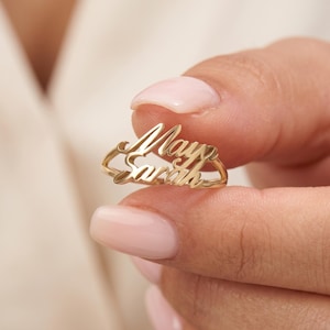Peut inclure: Une bague en métal doré présentant deux noms superposés en écriture cursive, May et Sarah, intégrés dans la structure de l'anneau. La bague possède une finition polie et réfléchissante ainsi qu'une structure ajourée délicate.