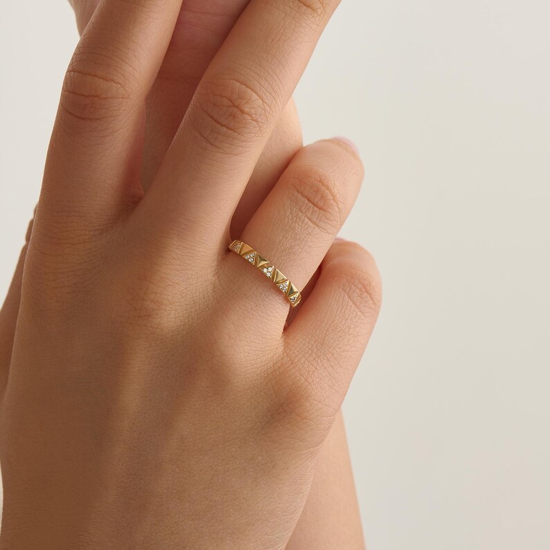 May include: A gold ring with a geometric pattern, set with small, clear gemstones. The ring is displayed on a finger, highlighting its detailed design and sparkling embellishments. The ring's warm gold hue contrasts with the skin, emphasising its elegance.
