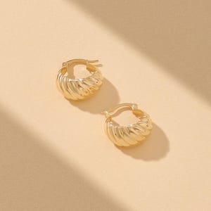 May include: A pair of gold-toned hoop earrings featuring a thick, rounded, and ribbed texture. The earrings have a hinged post closure mechanism and a polished metallic finish, showcasing a curved, croissant-like shape with distinct ridges.