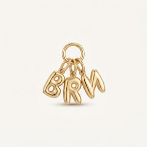 Könnte beinhalten: Ein goldfarbener Metallanhänger mit drei einzelnen, aufgeblähten Buchstabenanhängern, die an einem runden Sprungring hängen. Die Buchstaben B, R und M sind in einer glatten, abgerundeten, ballonartigen Schrift mit polierter Oberfläche.