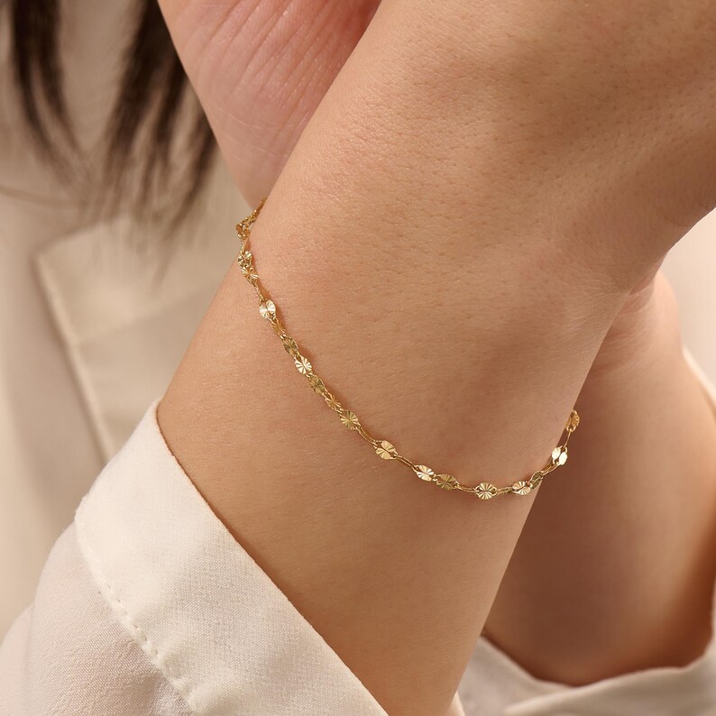 May include: A delicate gold chain bracelet featuring a series of small, circular, textured links that catch the light. The bracelet is thin and minimalist in its construction, consisting of repeating metallic discs connected by fine gold links.