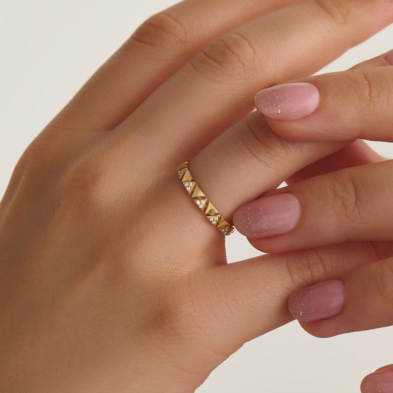 May include: A gold ring featuring a repeating pattern of triangular shapes, each set with small, clear gemstones. The ring is worn on a finger, highlighting its design and sparkle. The ring's design is geometric and modern.