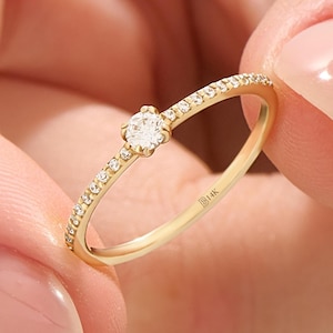 Könnte beinhalten: Ein zarter Goldring mit einem zentralen, runden Diamanten und kleineren Diamanten, die entlang des Bandes gesetzt sind. Der Ring ist mit "B14K" markiert.