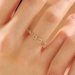 May include: A gold chain link ring featuring small, vibrant pink stones embedded along the center of each individual link. The ring has a delicate, repeating geometric design that encircles the finger, showcasing a polished metallic finish.