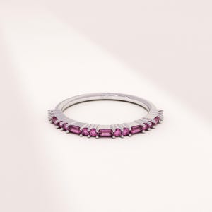 Op de afbeelding: Een zilveren ring met een band van kleine, rechthoekige, roze edelstenen. De edelstenen zijn gelijkmatig verdeeld over de ring, waardoor een doorlopende lijn van kleur ontstaat. De ring staat tegen een witte achtergrond.