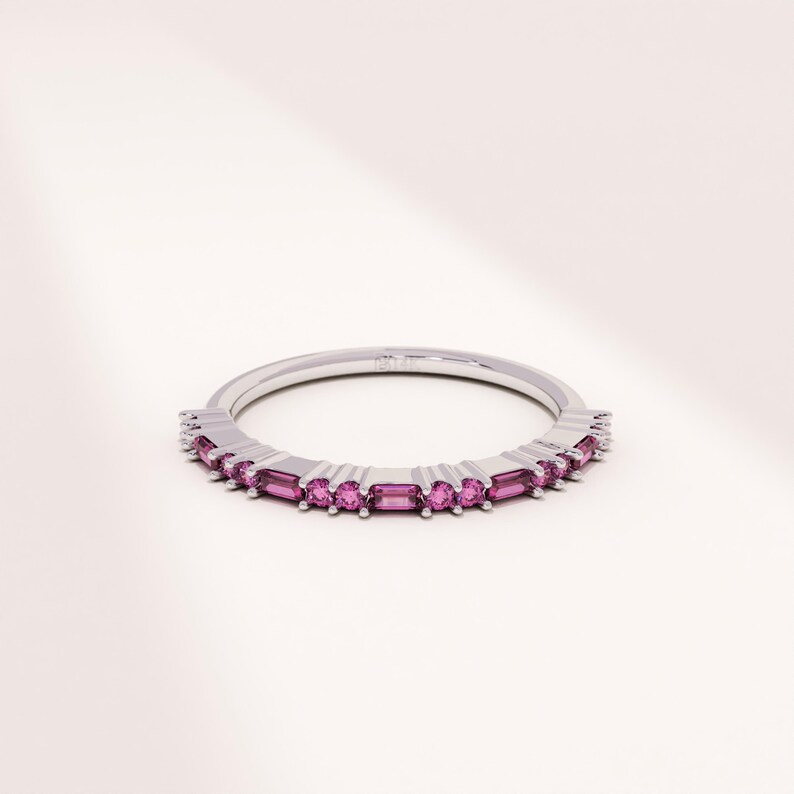 Op de afbeelding: Een zilveren ring met een band van kleine, rechthoekige, roze edelstenen. De edelstenen zijn gelijkmatig verdeeld over de ring, waardoor een doorlopende lijn van kleur ontstaat. De ring staat tegen een witte achtergrond.