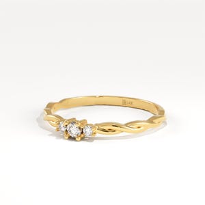 May include: A delicate gold ring with a braided band design. The ring features three small, clear gemstones clustered together. The ring is set against a white background, highlighting its intricate details.