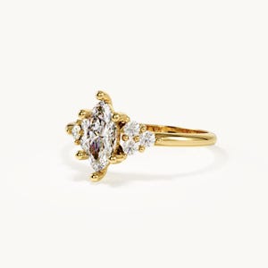 May include: A gold ring featuring a central marquise-cut diamond flanked by three smaller round-cut diamonds on each side. The band is polished yellow gold, and the stones are set in a prong setting, creating a cluster design on the ring face.