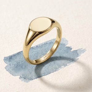 Op de afbeelding: Een gepolijste 14K gouden zegelring met een glad, ovaalvormig oppervlak. De binnenkant van de ring is duidelijk gegraveerd met de tekst 14K, wat de goudzuiverheid aangeeft. De ring heeft een solide, afgerond bandontwerp met een hoogglanzende metalen afwerking.