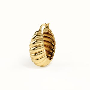 May include: A gold hoop earring featuring a thick, rounded, and ribbed croissant-like texture. The earring has a polished, reflective metallic finish and a hinged closure mechanism at the top, showcasing a curved, chunky, and dimensional design.