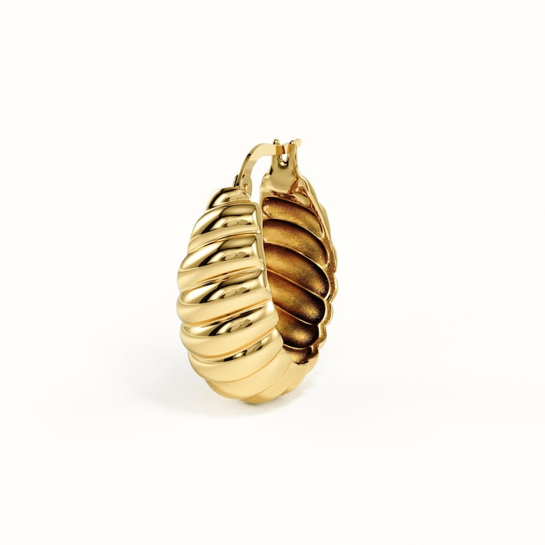 May include: A gold hoop earring featuring a thick, rounded, and ribbed croissant-like texture. The earring has a polished, reflective metallic finish and a hinged closure mechanism at the top, showcasing a curved, chunky, and dimensional design.