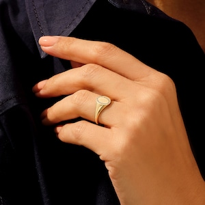 May include: A gold signet ring with a round, faceted stone set in the center. The ring is worn on the index finger of a hand.
