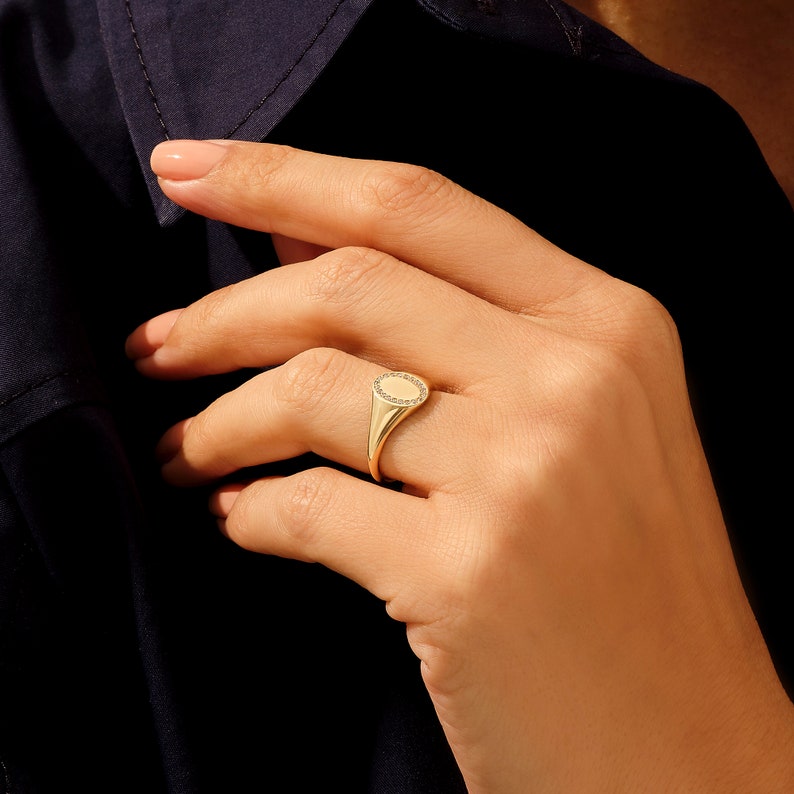 May include: A gold signet ring with a round, faceted stone set in the center. The ring is worn on the index finger of a hand.