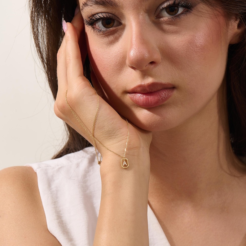 May include: A fine gold necklace with a pendant displaying the letter "A" within a square. The chain is slender, and the pendant is small, offering a minimalist and elegant aesthetic. The necklace is shown against a white backdrop.