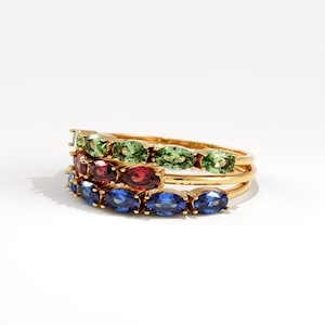 May include: Three gold rings stacked together, each with a row of oval-cut gemstones. The top ring has green stones, the middle ring has red stones, and the bottom ring has blue stones.