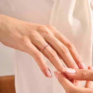 Puede incluir: Un anillo de oro con una banda de peque&ntilde;as piedras preciosas de color rosa vibrante. El anillo est&aacute; en el dedo de una persona, con la mano de otra persona cerca. El fondo es una tela suave de color blanquecino.