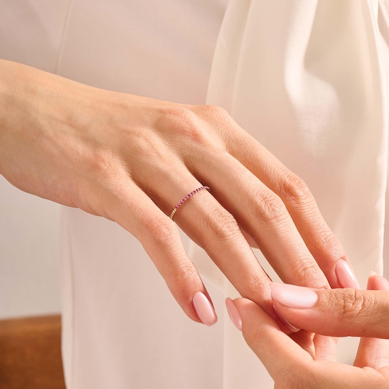 Puede incluir: Un anillo de oro con una banda de peque&ntilde;as piedras preciosas de color rosa vibrante. El anillo est&aacute; en el dedo de una persona, con la mano de otra persona cerca. El fondo es una tela suave de color blanquecino.
