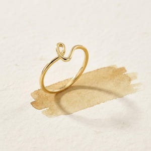 May include: A polished gold ring featuring a thin, continuous band that curves upward into a small, open loop design at the top. The metal has a smooth, reflective surface, showcasing a simple, minimalist structure with a rounded profile.